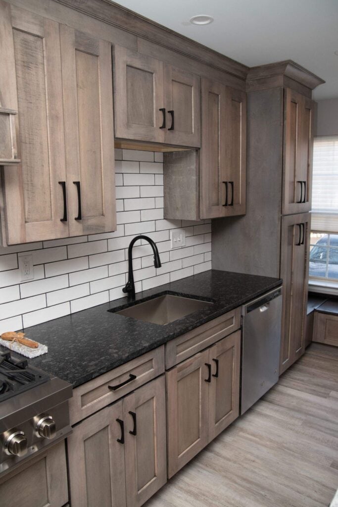 Modern kitchen with light wood cabinets, black handles, black countertop, stainless steel sink with black faucet, white subway tile backsplash, stainless steel dishwasher, and a gas stove. Natural light fills the room.
