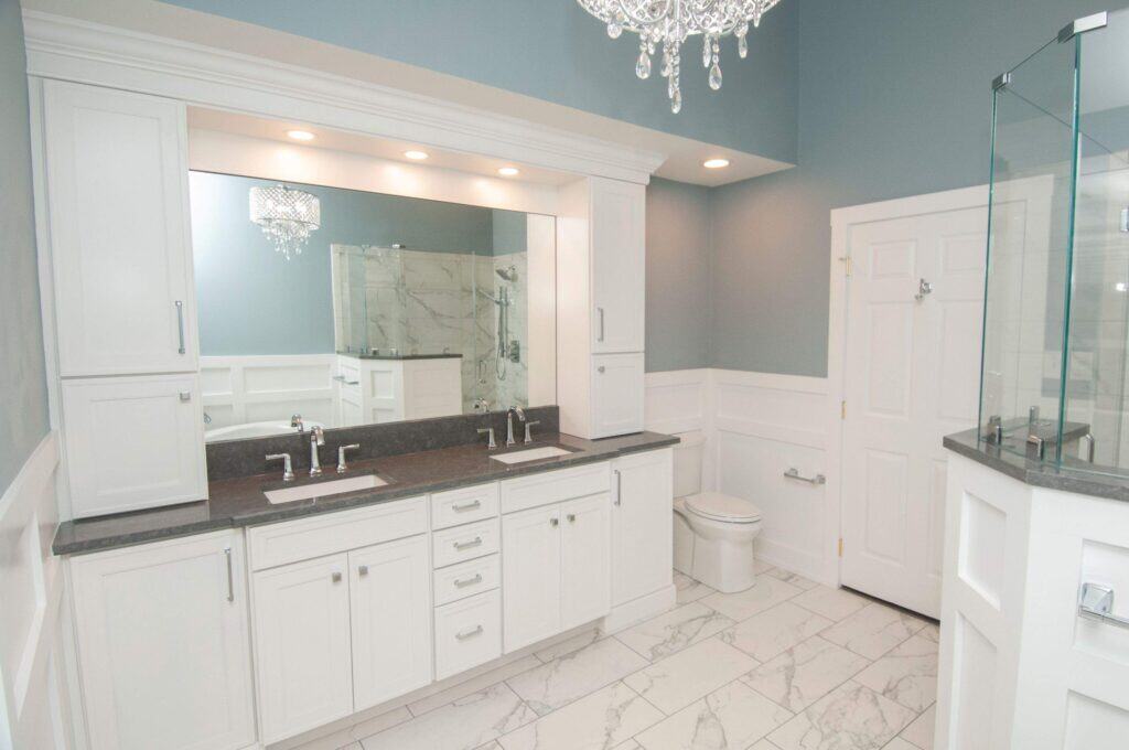 Modern bathroom with double sinks, white cabinets, gray countertops, a large mirror, a glass shower, a toilet, marble floor tiles, and a chandelier, all in a light and airy color scheme.