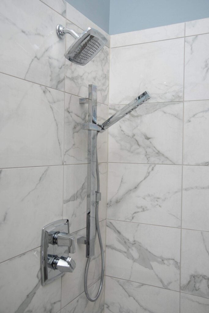 Modern shower with marble-tiled walls, featuring a square overhead showerhead, a handheld shower attachment, and chrome controls mounted on the wall.