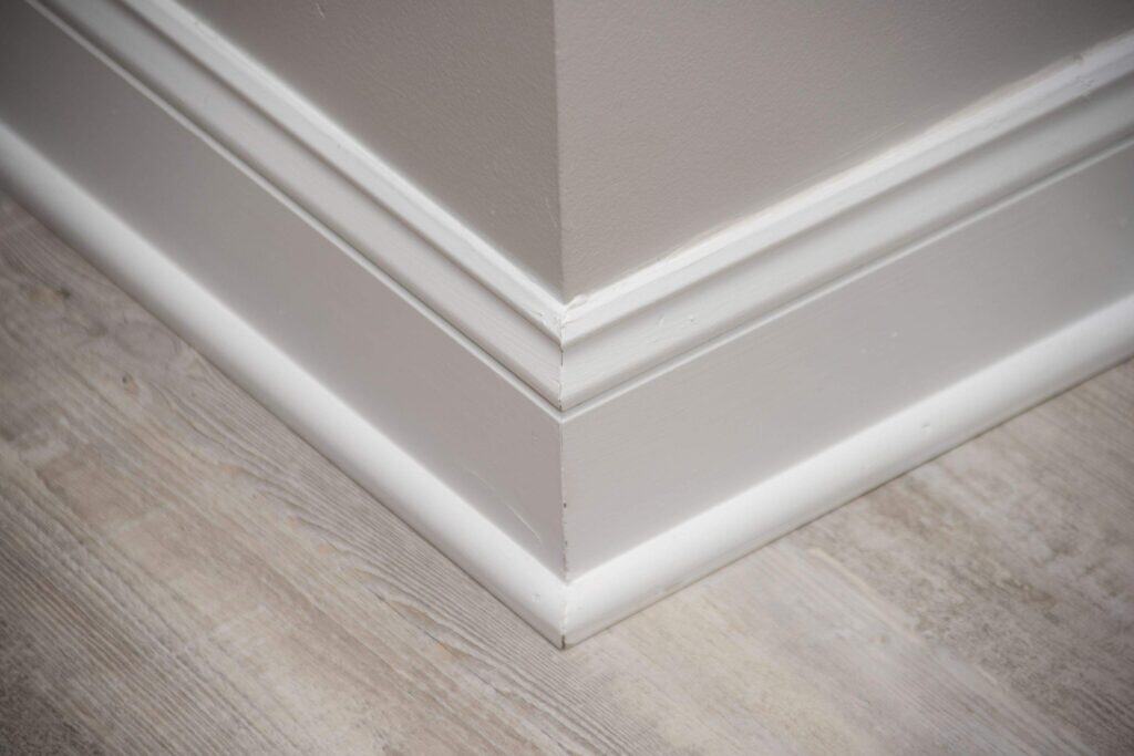 Close-up view of a white baseboard molding at the corner of a wall, with light-colored wood flooring. The corner is neatly joined, showing clean lines and detailed trim work.