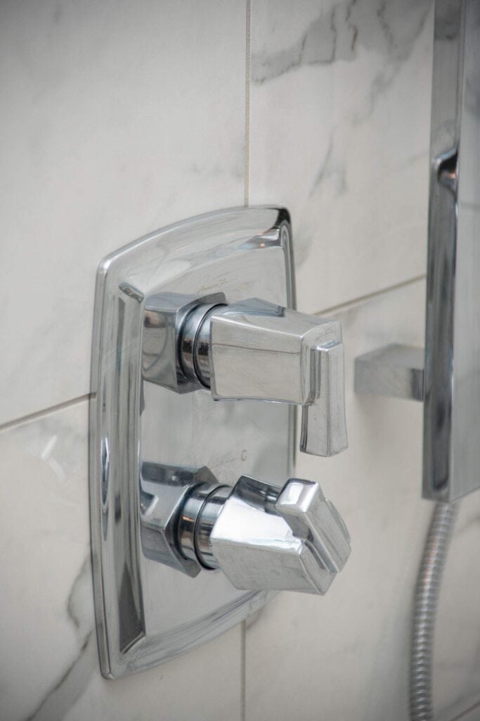 Close-up of a modern chrome shower control panel with two angular knobs mounted on a marble-tiled wall; one likely controls water flow, the other adjusts temperature.