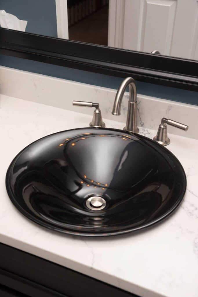 A modern bathroom sink with a glossy black bowl, silver faucet, and handles, set in a white marble countertop with a black-framed mirror in the background.