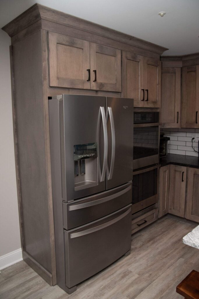A modern stainless steel French door refrigerator with a bottom freezer, built into wood cabinetry in a kitchen with light wood floors and dark countertops.