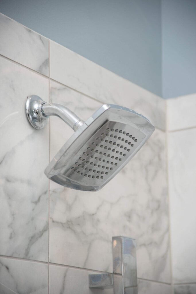 A close-up view of a modern, square-shaped chrome showerhead mounted on a tiled bathroom wall with a marble pattern.