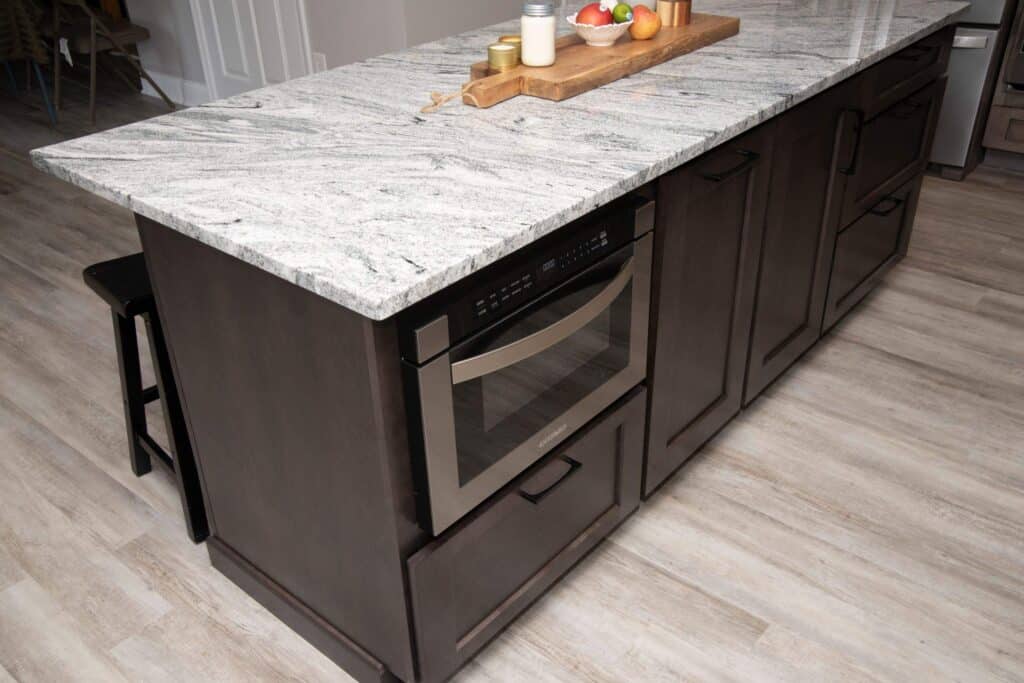 A modern kitchen island with dark wood cabinets, a built-in microwave, a light-colored granite countertop, and a wooden tray holding fruit, milk, and spices. The island sits on light wood flooring.