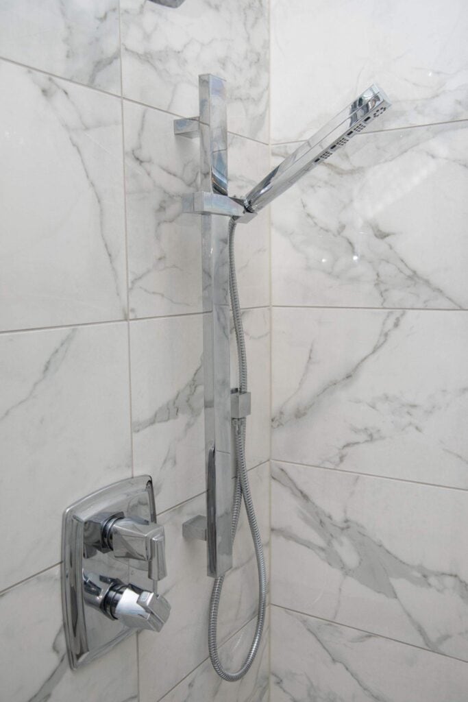 Modern chrome shower fixture with a handheld showerhead attached to a hose, mounted on a wall with white and gray marble tiles. Two square temperature and flow control knobs are also visible on the wall.