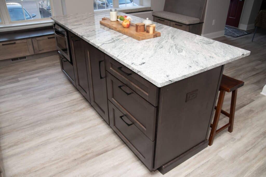 A modern kitchen island with a white and gray marble countertop, dark brown cabinets, black handles, and a wooden barstool. The countertop has a wooden tray with candles, apples, and a jar. Light wood flooring surrounds the island.