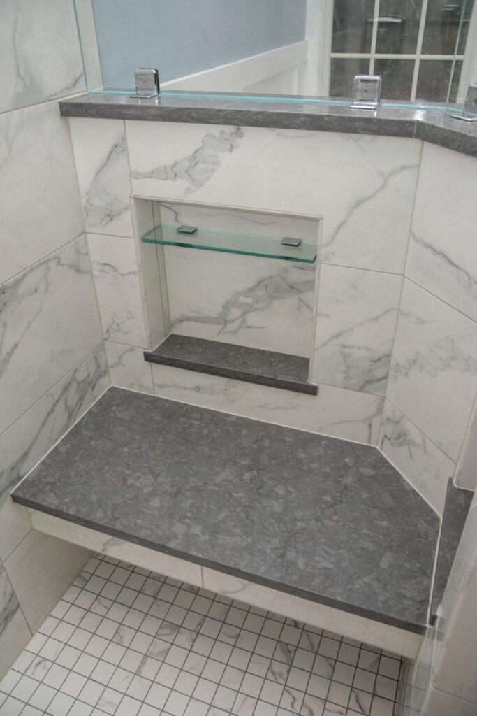 Marble-tiled shower with a built-in gray bench, a glass shelf, and a small ledge below it; the floor is covered with small white square tiles.