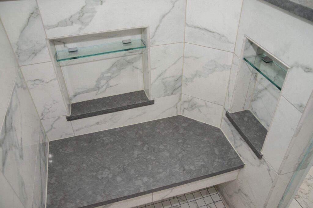 A shower corner with a built-in gray stone bench, a matching gray shelf, glass shelf, and a small recessed mirror, all set against white marble-patterned tiles.