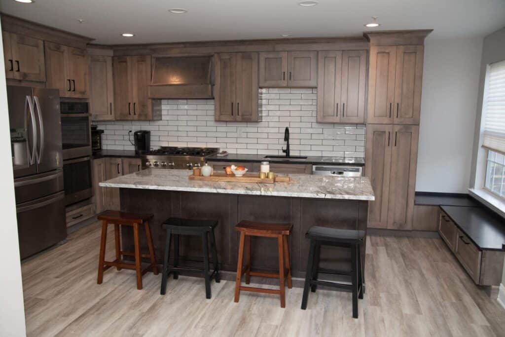 Modern kitchen with wood cabinets, stainless steel appliances, white subway tile backsplash, marble island countertop, and four stools. Light wood flooring and large window with bench seating are also visible.