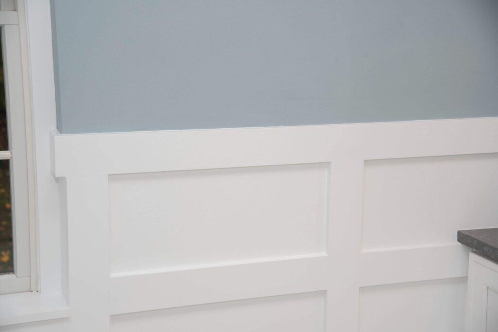 A section of wall with white board and batten wainscoting and a light blue upper wall, next to a window with white trim and part of a dark countertop visible on the right.