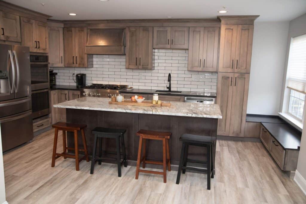 Modern kitchen with light wood cabinets, stainless steel appliances, white subway tile backsplash, marble island countertop, and four mismatched stools. There’s a window seat with storage on the right.