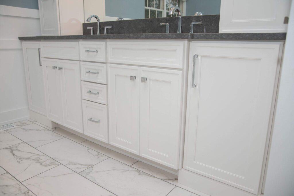 White bathroom vanity with multiple drawers and cabinets, silver handles, black countertop, and double sinks. The floor features large white tiles with gray veining, and the vanity is set against a light blue wall.