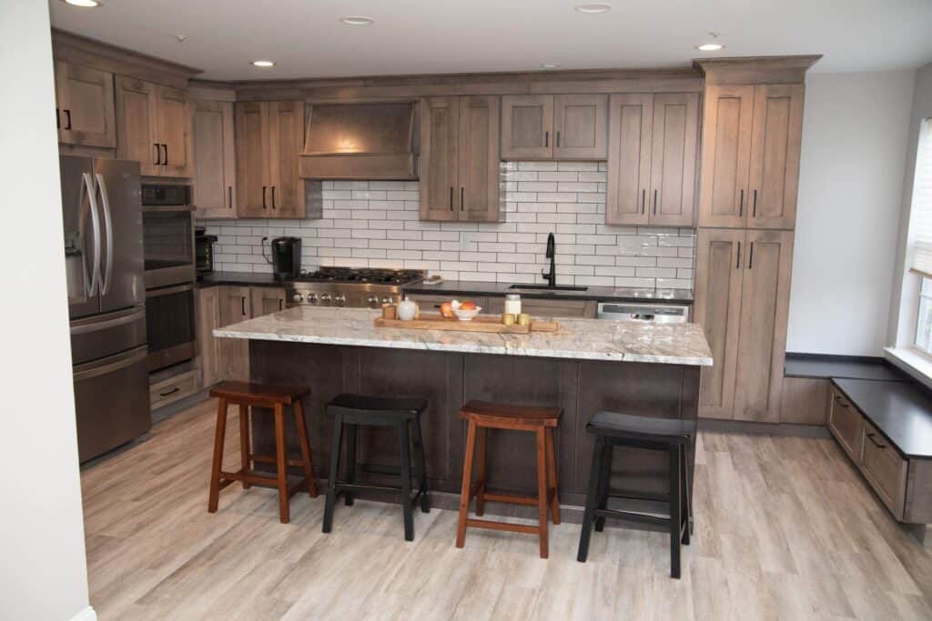 Modern kitchen with wood cabinets, stainless steel appliances, a white subway tile backsplash, and a large island with marble countertop. Four wooden stools are placed at the island, and there is a window seat on the right.