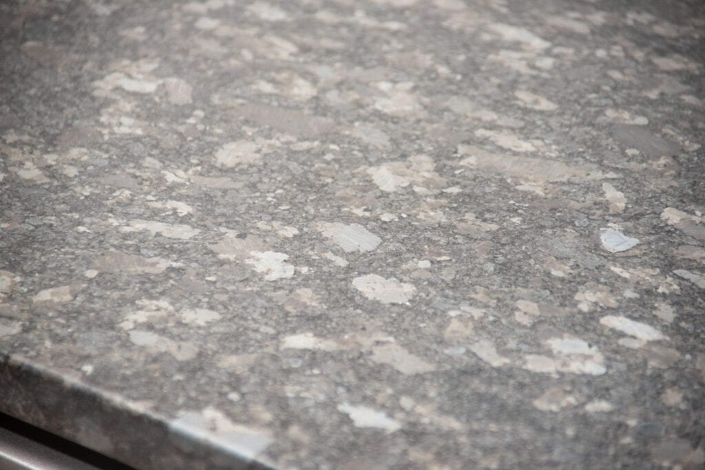 Close-up of a gray granite countertop with a polished surface, showing natural patterns of lighter and darker speckles and patches. The edge of the countertop is visible in the lower left corner.
