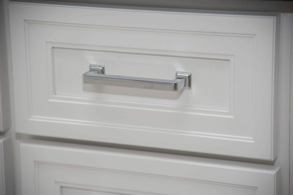A close-up of a white cabinet drawer with a rectangular silver handle. The drawer features raised panel detailing and is part of a larger cabinet.