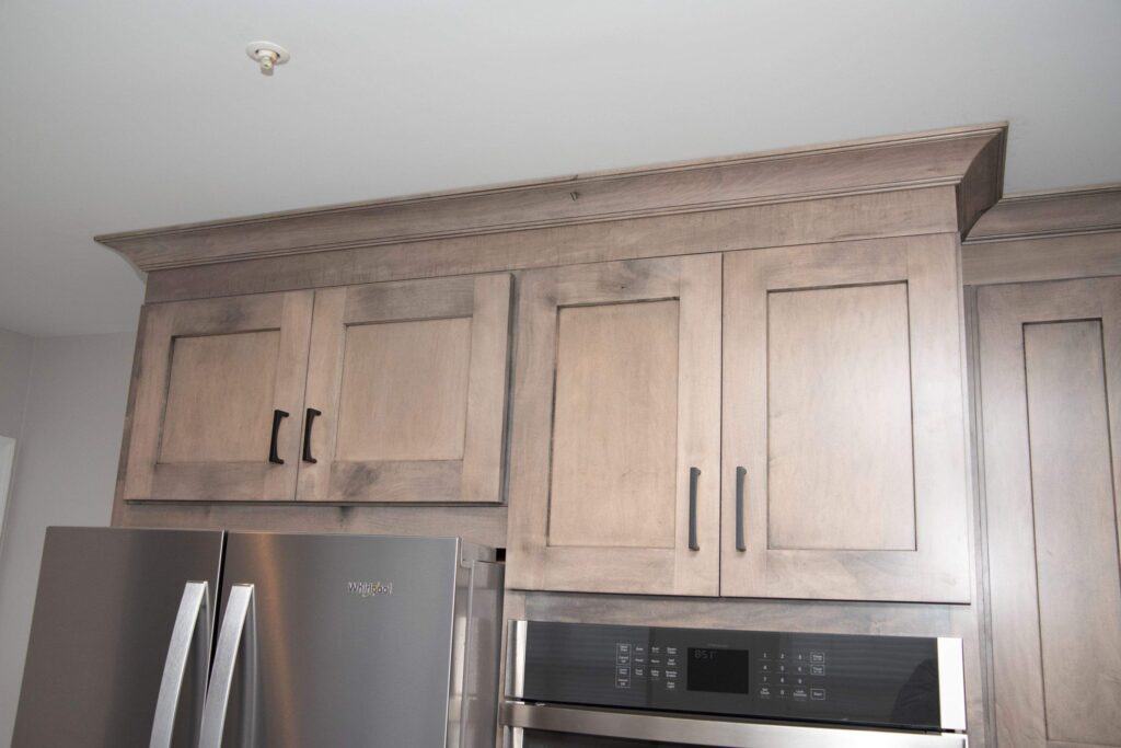 Upper kitchen cabinets with a natural wood finish, black handles, a stainless steel refrigerator, and a built-in double oven underneath. The cabinets extend up to a crown molding near the ceiling.