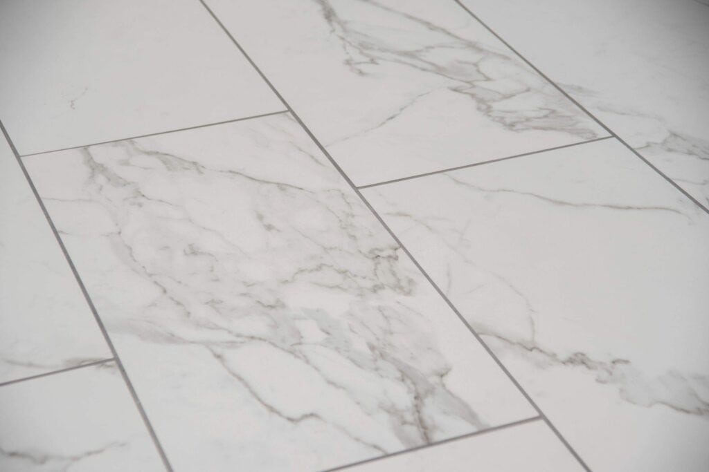 Close-up of white marble floor tiles with grey veins and light grout lines, arranged in a staggered pattern. The surface appears smooth and polished.