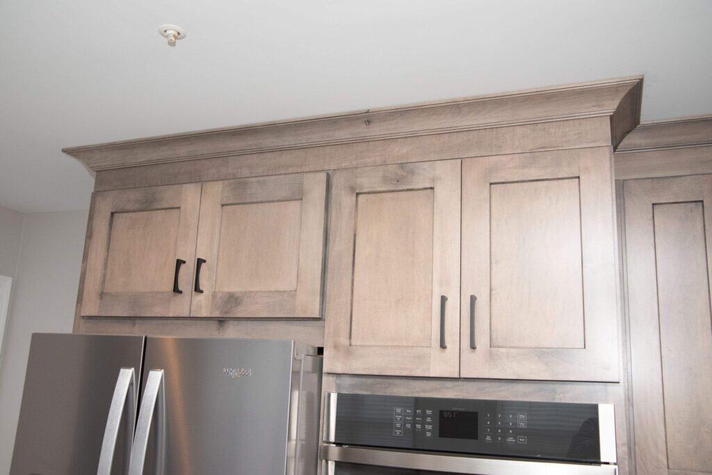 Light wood kitchen cabinets with black handles are installed above a stainless steel refrigerator and a built-in oven. The cabinets feature simple panel doors and crown molding at the top.