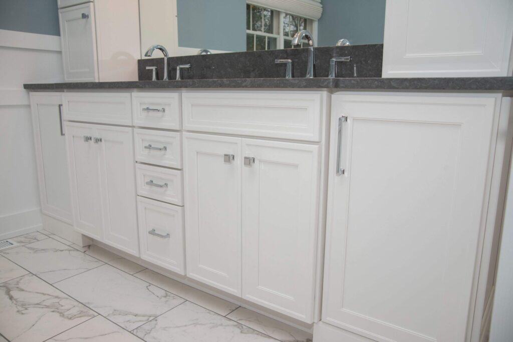 White bathroom vanity with double sinks, dark countertop, chrome faucets, and multiple cabinet doors and drawers. The vanity sits on a white marble tile floor with gray veining. A mirror and window are partially visible.