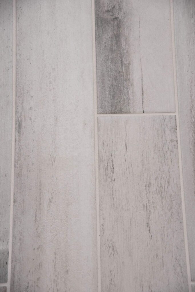Close-up of light gray and white rectangular tiles with a wood grain pattern, arranged in a staggered layout. The surface appears textured, resembling weathered or distressed wood planks.