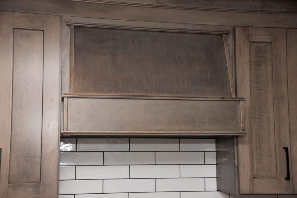 Wooden kitchen cabinets with a range hood cover above a white subway tile backsplash. The cabinets and hood have a natural wood finish and simple, clean lines.
