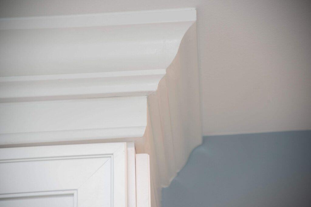Close-up of white crown molding at the junction of a wall and ceiling, with part of a cabinet visible below. The wall is painted blue on the lower part and light gray above.