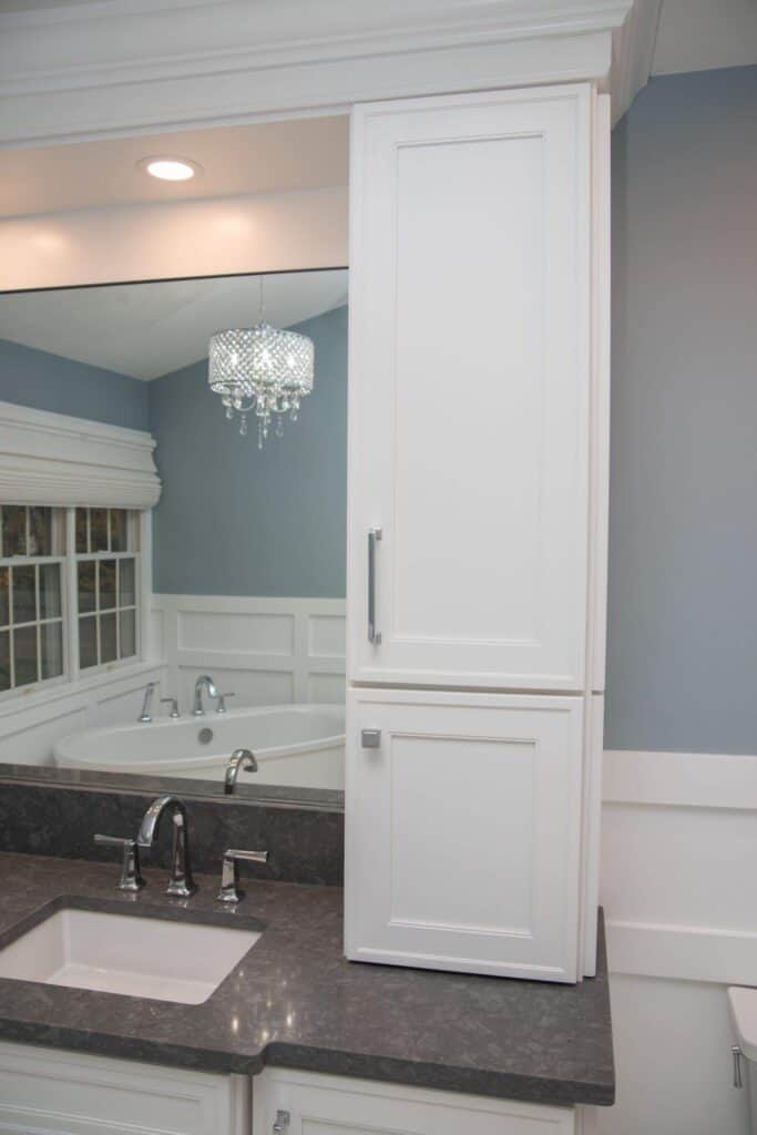 A bathroom with a white cabinet, gray countertop, and an under-mount sink. A chrome faucet is set in front of a large mirror, and a small chandelier hangs from the ceiling. The walls are painted light blue.