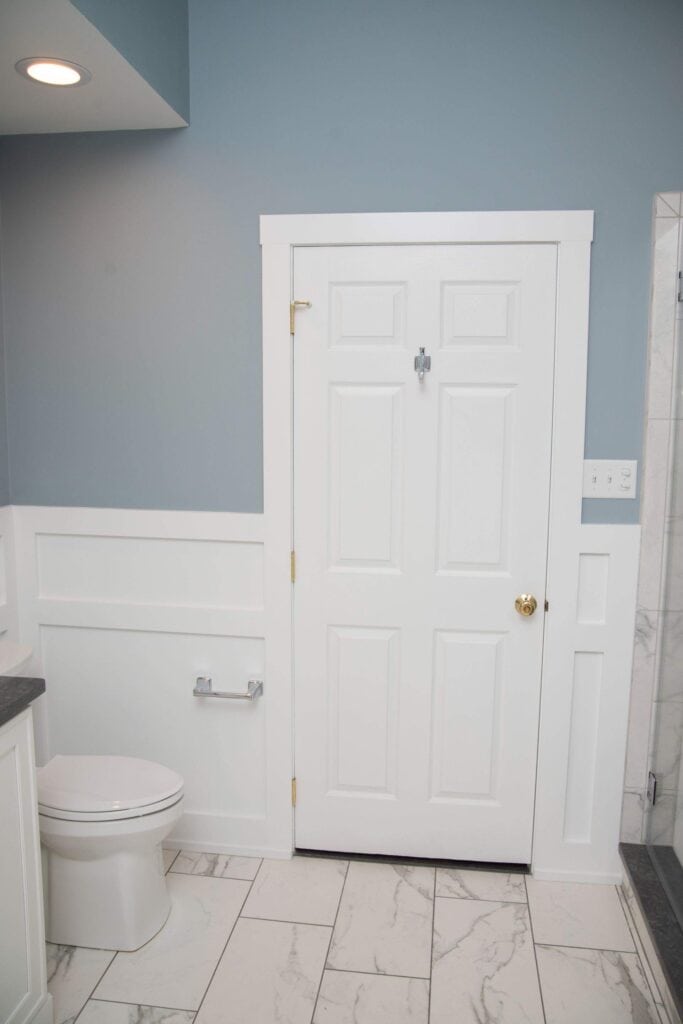 A modern bathroom with light blue walls, white wainscoting, a white door, a white toilet, chrome toilet paper holder, and white tile floor with grey veining. A shower with a glass door is partially visible on the right.