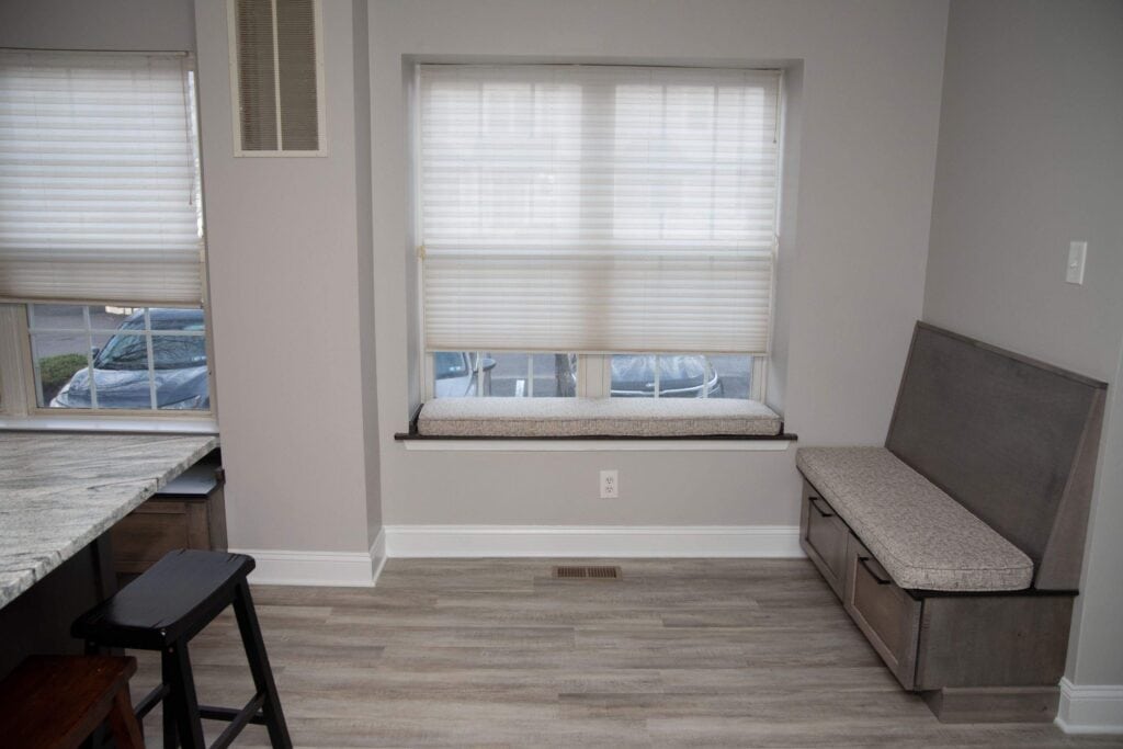 A small, modern room with light gray walls and flooring, featuring a window seat with a cushion under a large window, a built-in storage bench with cushion, and part of a marble countertop and black stool visible.
