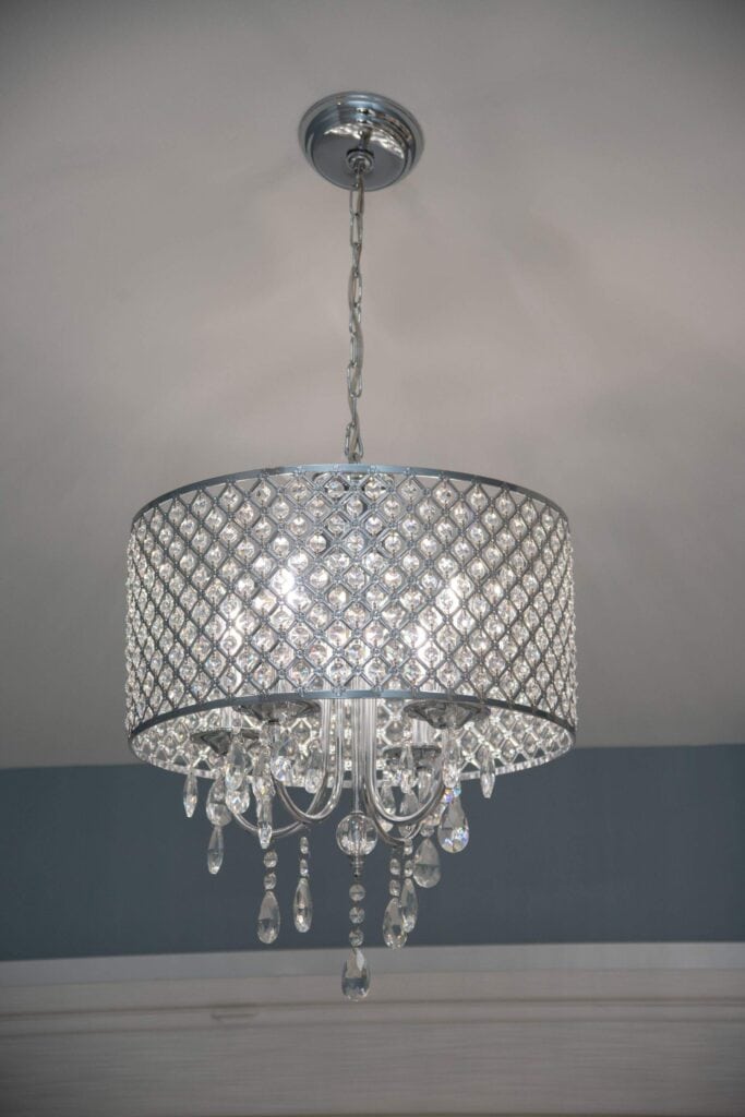 A modern crystal chandelier with a silver chain and drum-shaped shade featuring a diamond pattern, adorned with hanging crystal pendants, is lit and mounted on a ceiling.