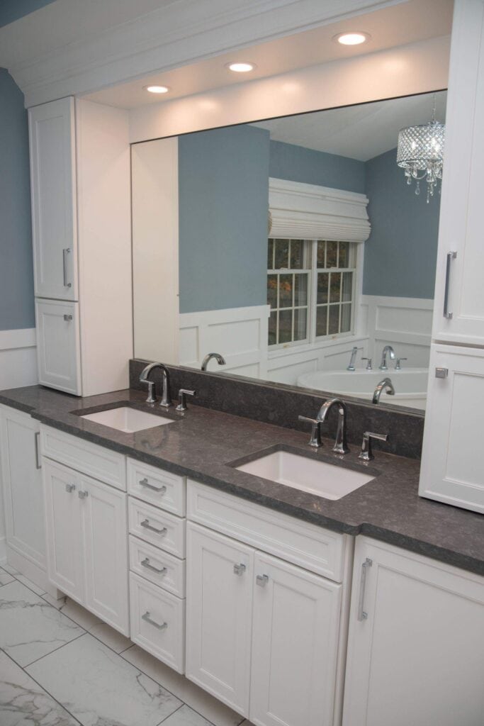 A modern bathroom with two sinks, white cabinets, a dark countertop, large mirror, recessed lighting, blue walls, and a window with white trim reflected in the mirror.