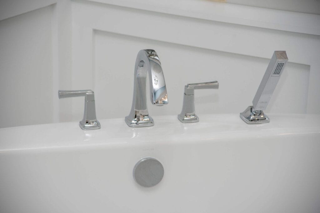 Close-up of a modern bathtub with chrome fixtures, including a faucet, two handles, and a handheld showerhead, all mounted on the edge of a white tub against a white paneled wall.