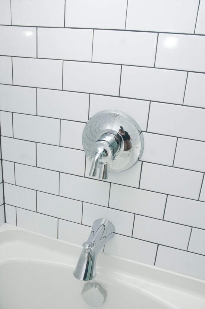 Close-up of a chrome shower faucet and spout installed on a white subway tile wall above a bathtub. The tiles are arranged in a horizontal pattern with visible grout lines.