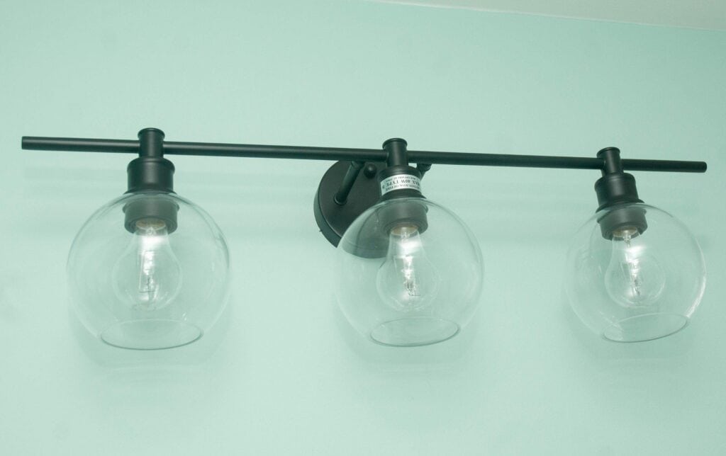 A black, wall-mounted light fixture with three clear glass globe shades, each containing a visible light bulb, set against a light green wall.