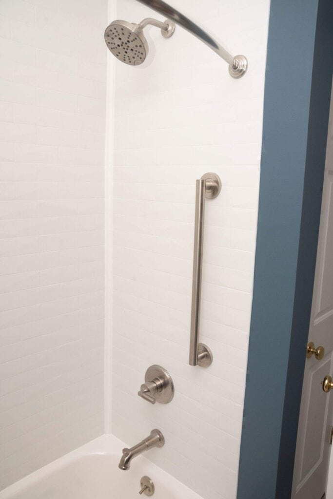 A modern bathtub and shower combo with white tile walls, a silver showerhead, faucet, and a long vertical grab bar for safety. The edge of a blue wall and a white door are visible on the right.