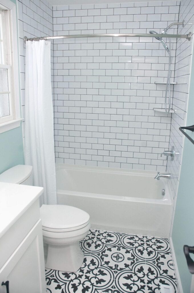 A bathroom with white subway tile walls, a white bathtub with a showerhead and curtain, a white toilet, a white vanity, and black-and-white patterned floor tiles. Light blue walls and a window are also visible.
