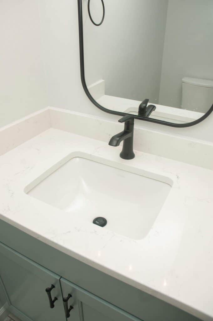 A bathroom vanity with a white marble countertop, an undermount rectangular sink, a matte black faucet, and matching black hardware. A portion of a black-framed mirror and a toilet are visible in the background.