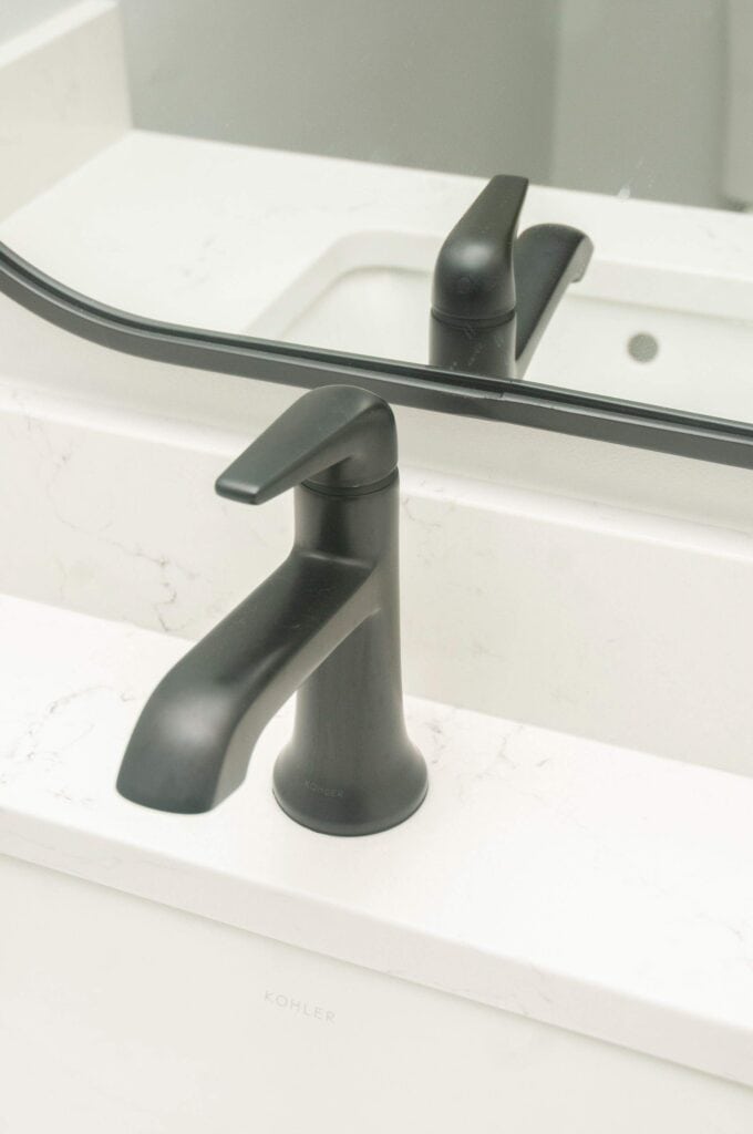 A modern black bathroom faucet is mounted on a white marble countertop in front of a rectangular mirror. The sink and part of the wall are also visible in the background.