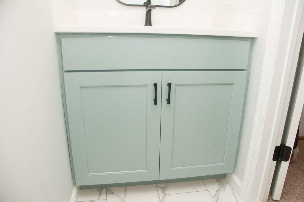 Light green bathroom vanity with two cabinet doors and black handles, set against a white wall and white marble tile floor. The vanity has a simple, modern design with a white countertop.
