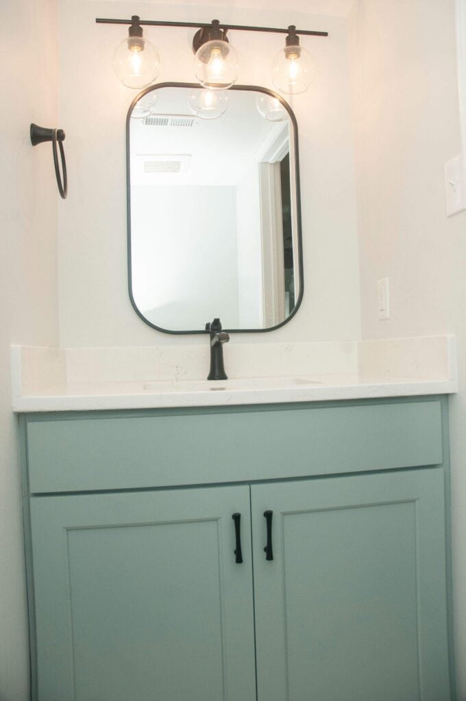 A modern bathroom vanity with light green cabinets, a white countertop, a black faucet, and a square mirror. Above the mirror are three globe light bulbs on a black fixture. The walls are painted white.