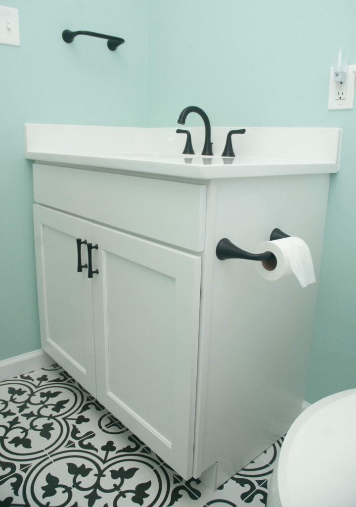White bathroom vanity with black hardware and faucet, mounted against a light blue wall. The cabinet has a toilet paper holder attached to the side and is set on a black and white patterned tile floor.