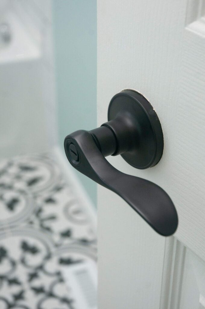 A close-up of a matte black lever door handle on a white door, with part of a patterned black and white tile floor visible in the background.