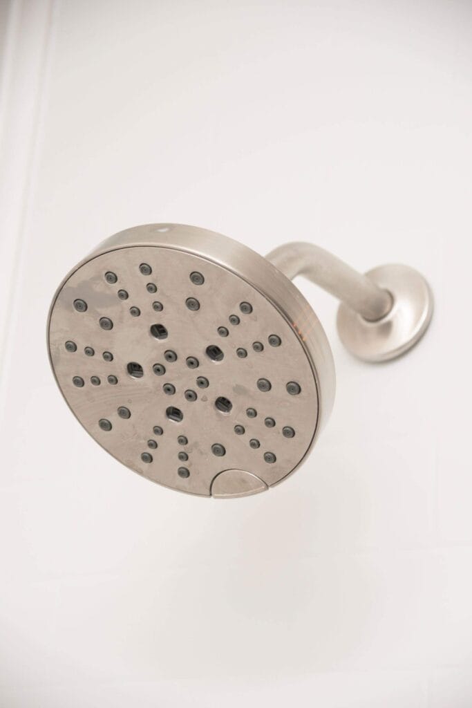 A close-up view of a modern, round, stainless steel showerhead attached to a wall, photographed from below against a white tiled background.