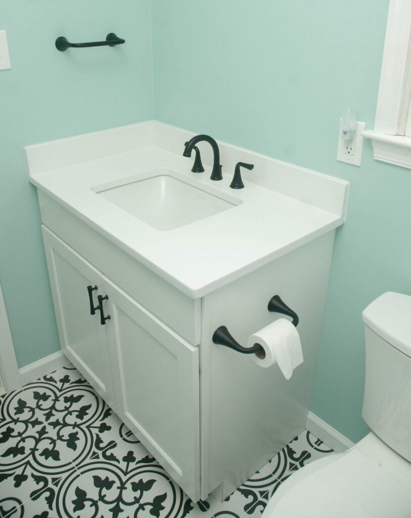 A small bathroom with a white vanity, black faucet, toilet paper holder, and towel bar, mint green walls, and black-and-white patterned tile floor. The edge of a toilet is visible on the right.