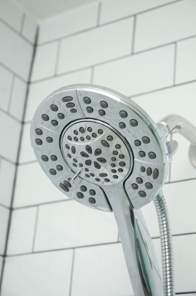 Close-up of a modern, silver showerhead with multiple spray settings, mounted on a wall with white subway tiles and gray grout in a bathroom.