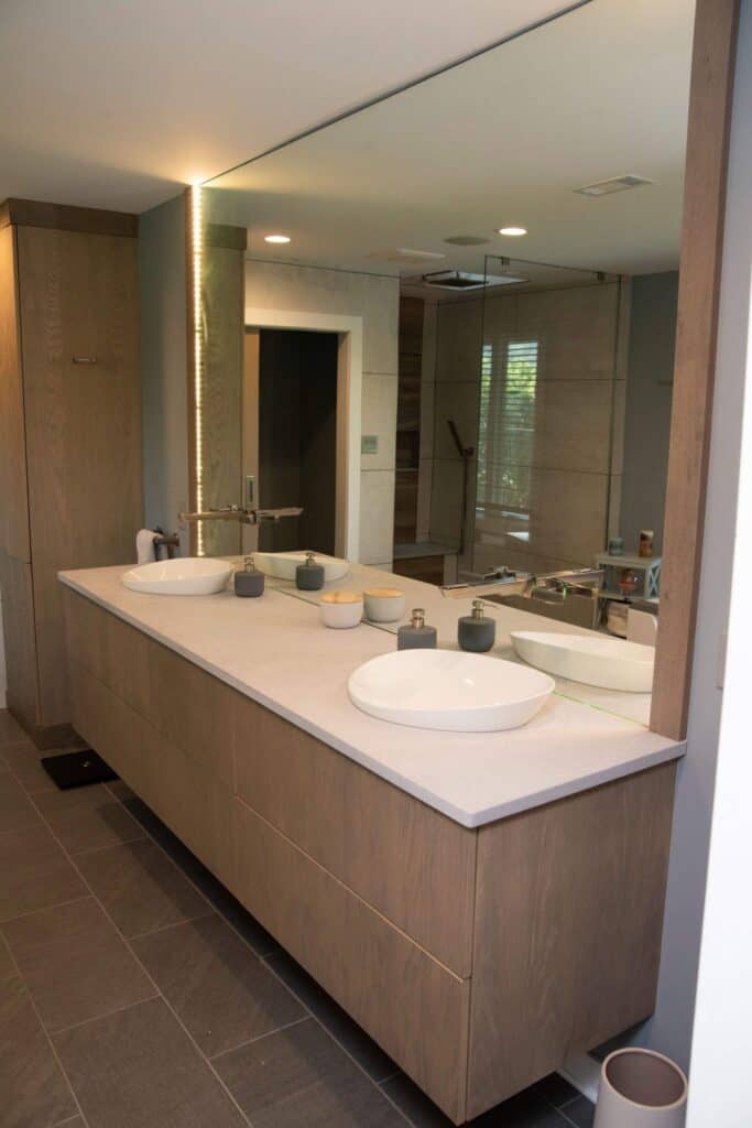 A modern bathroom with a large mirror, two white vessel sinks on a wooden floating vanity, and minimalistic decor. Glass shower enclosure and neutral tones create a clean, contemporary feel.