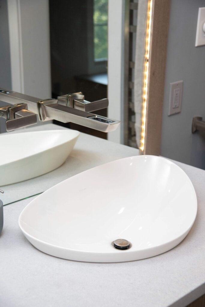 Modern bathroom sink with an asymmetrical, oval-shaped white basin on a light countertop. A sleek, rectangular chrome faucet is mounted above the sink, and a large mirror is visible in the background.