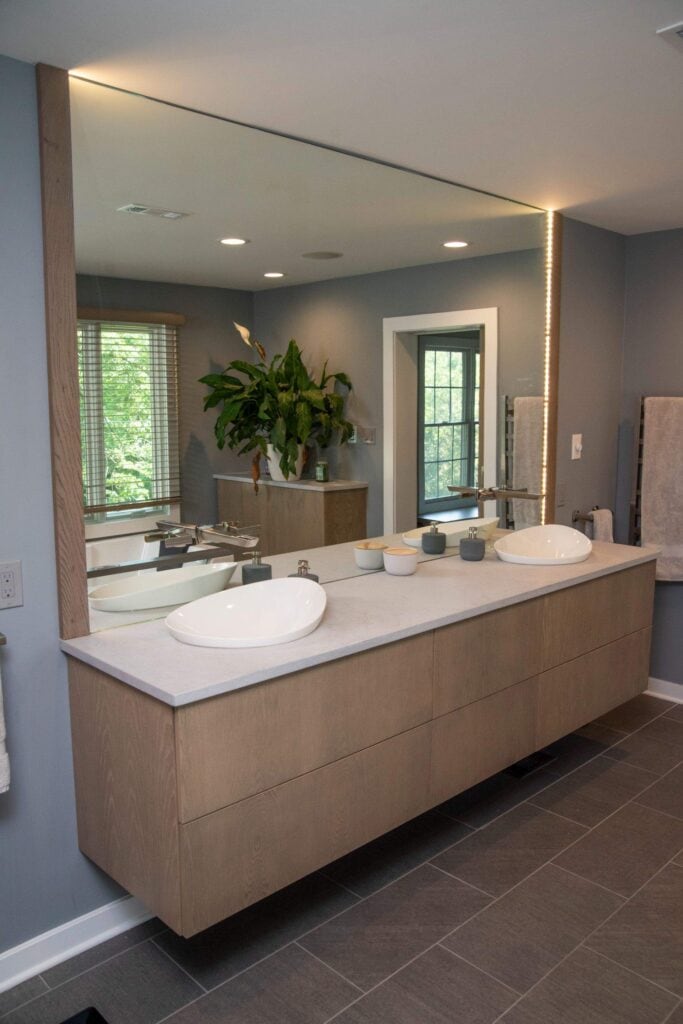 Modern bathroom with a floating wood vanity, two vessel sinks, a large mirror with built-in LED lighting, gray tiled floor, and a potted plant by the window in the background.