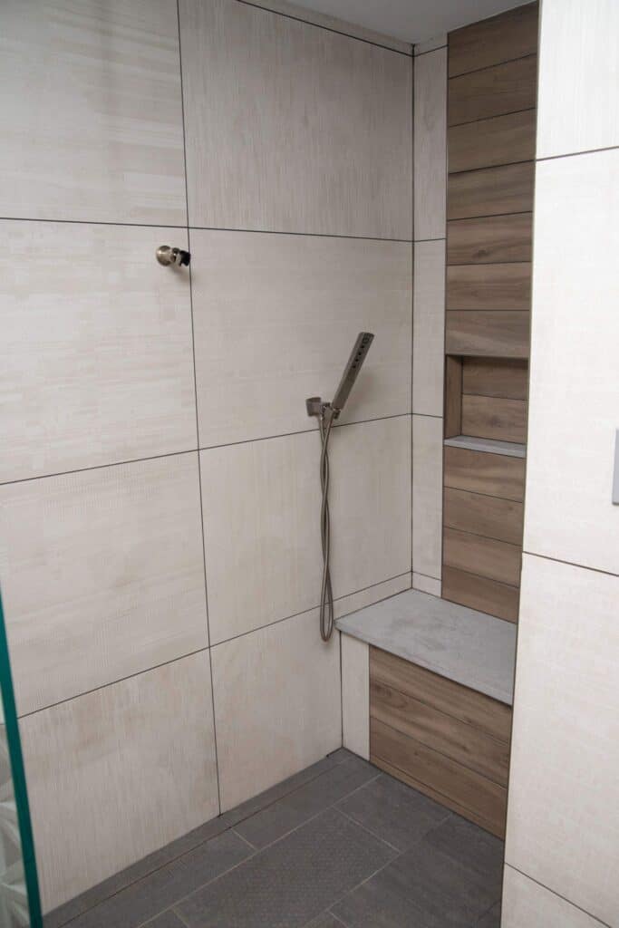 A modern shower with light beige tiles, a handheld showerhead attached to the wall, a built-in wood-look tiled bench, and matching recessed shelves.